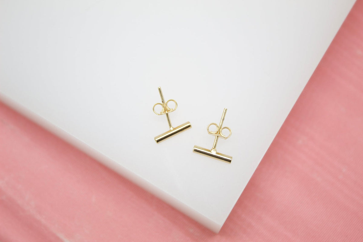 A pair of minimalist gold bar stud earrings rests on a white display surface against a soft pink background.