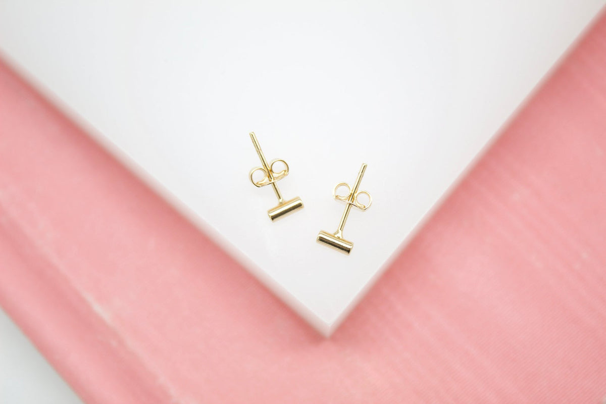 A pair of minimalist gold bar stud earrings rests on a white and pink display surface.