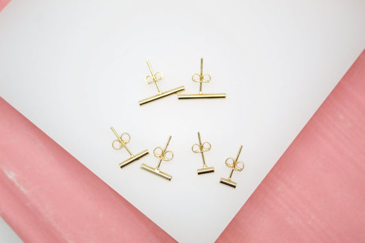 Several pairs of minimalist gold bar stud earrings rest on a white surface against a pink background.