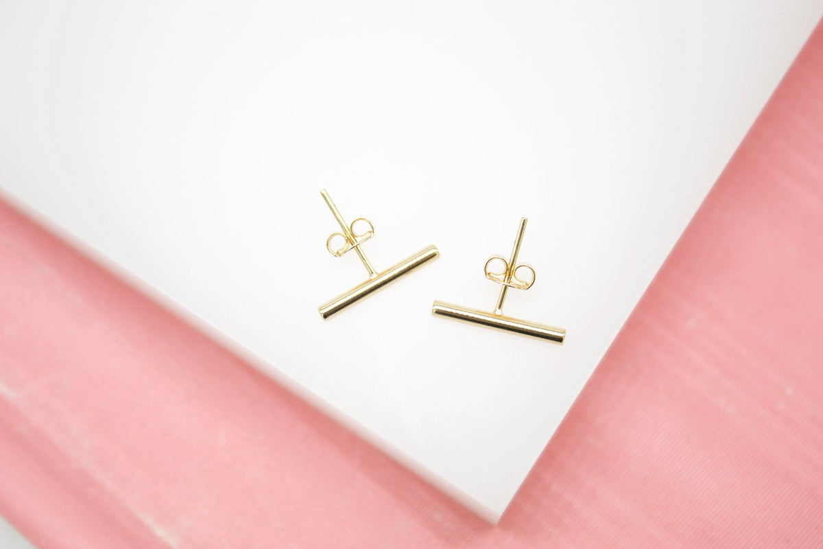 A pair of minimalist gold bar stud earrings rests on a white display surface with a pink background.