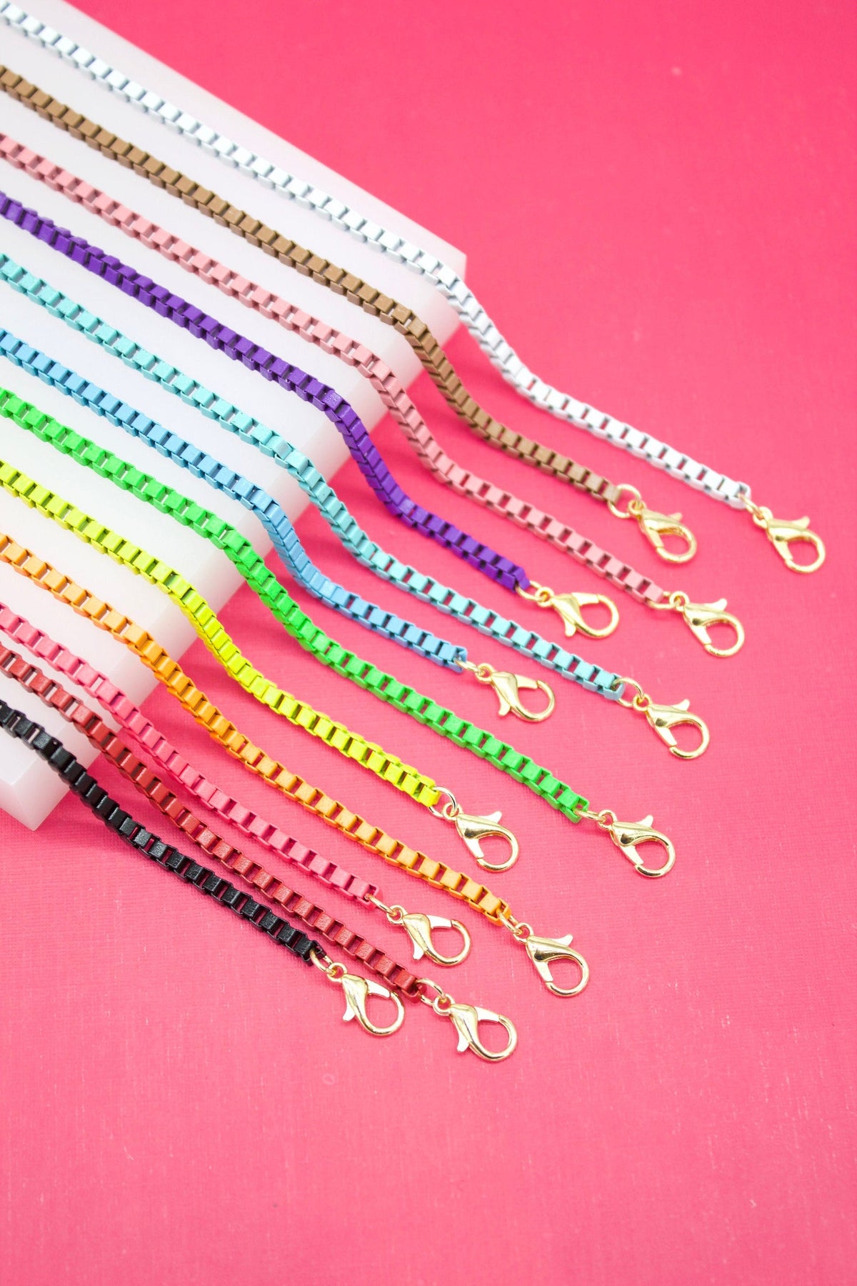 A vibrant collection of colorful enameled box chain necklaces with gold-toned lobster clasps rests on a white display block against a bright pink background.
