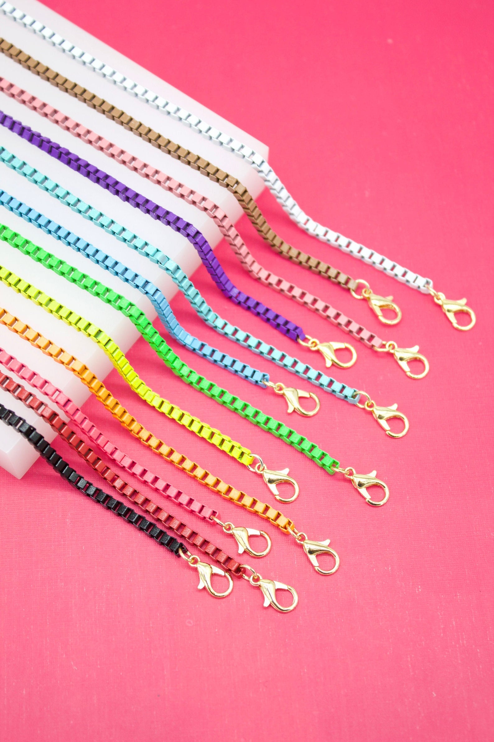 A vibrant collection of colorful enameled box chain necklaces with gold-toned lobster clasps rests on a white display block against a bright pink background.