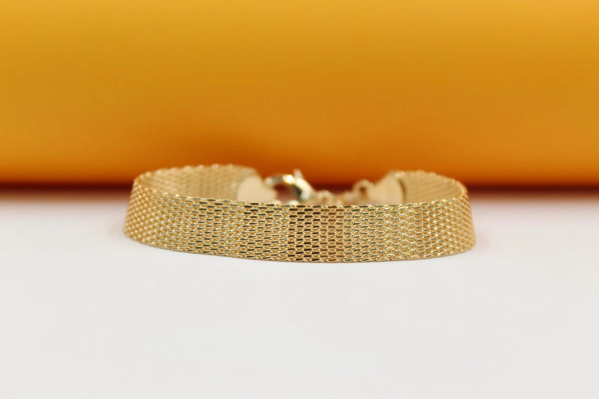 A wide gold mesh bracelet rests on a smooth surface against a warm yellow and white background.
