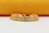 A wide gold mesh bracelet rests on a smooth surface against a warm yellow and white background.