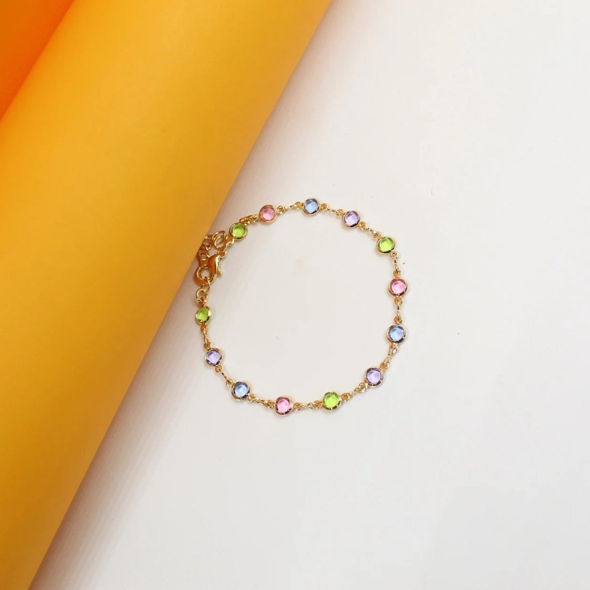 A dainty gold chain bracelet featuring multi-colored bezel-set crystals lies on a curved white and orange background.