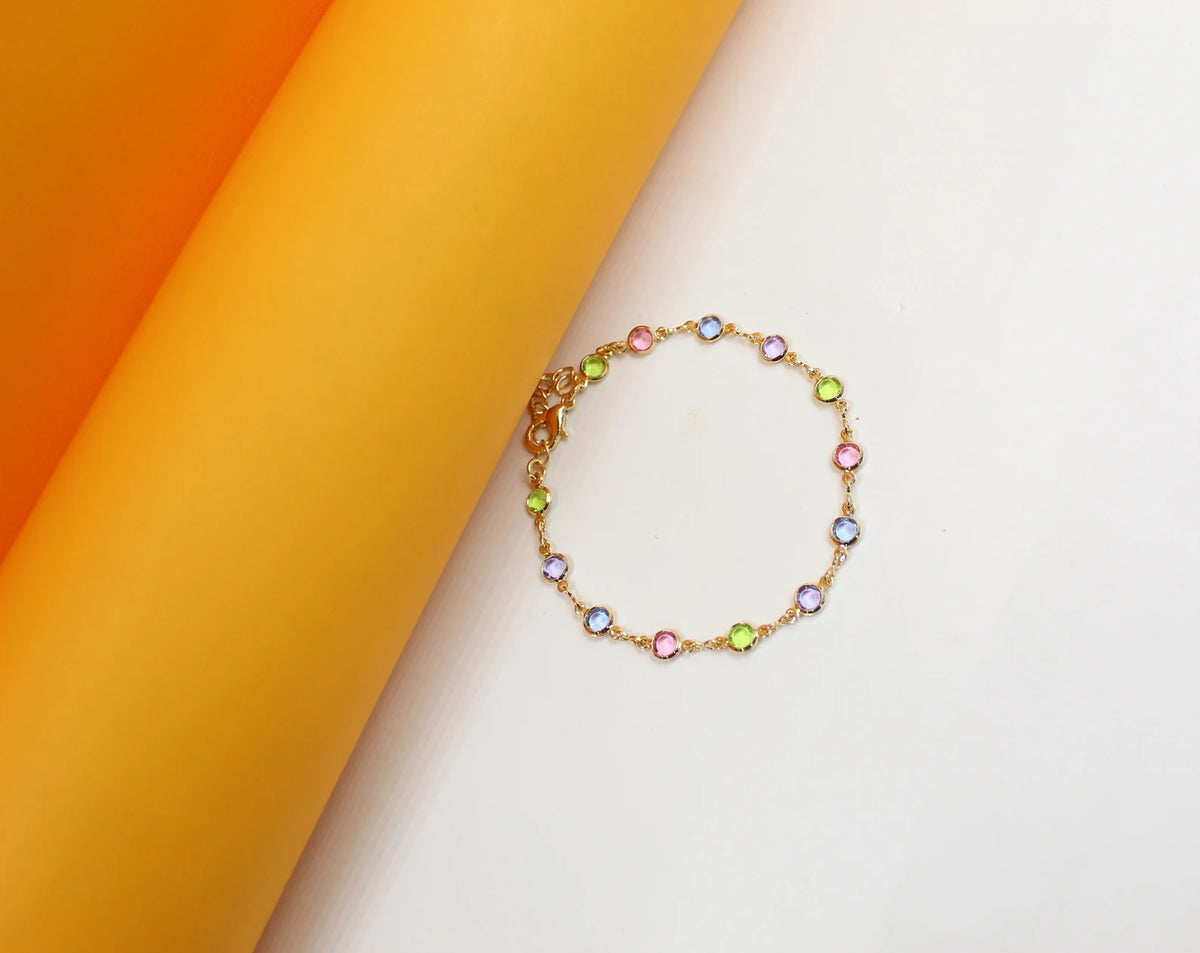 A delicate gold chain bracelet featuring colorful, bezel-set cubic zirconia stones rests against a vibrant yellow and white background.