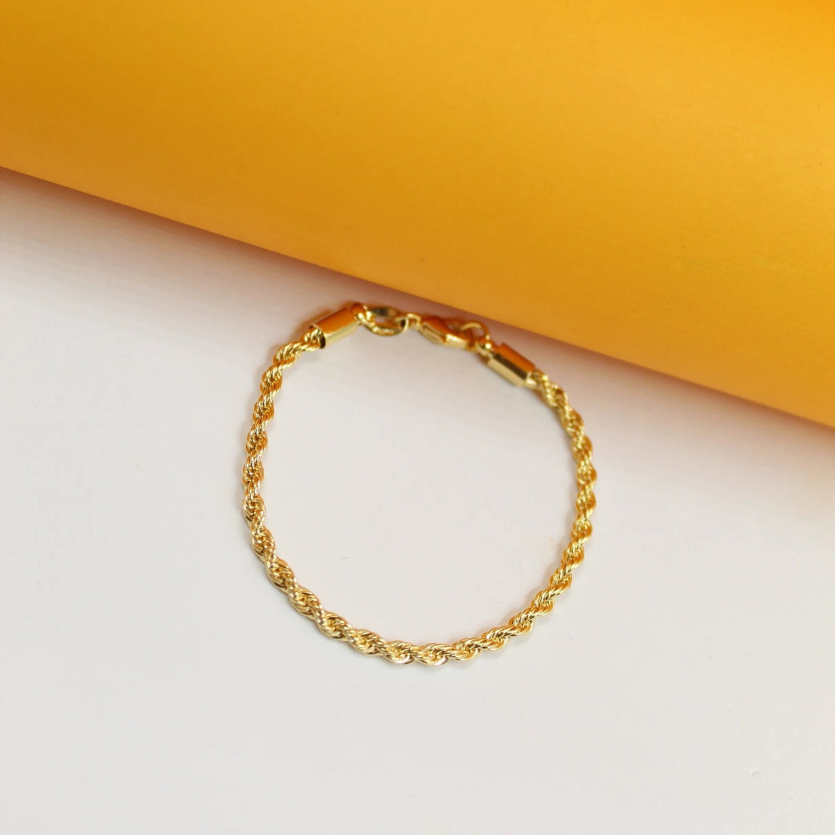A delicate gold rope chain bracelet rests on a white and yellow background.