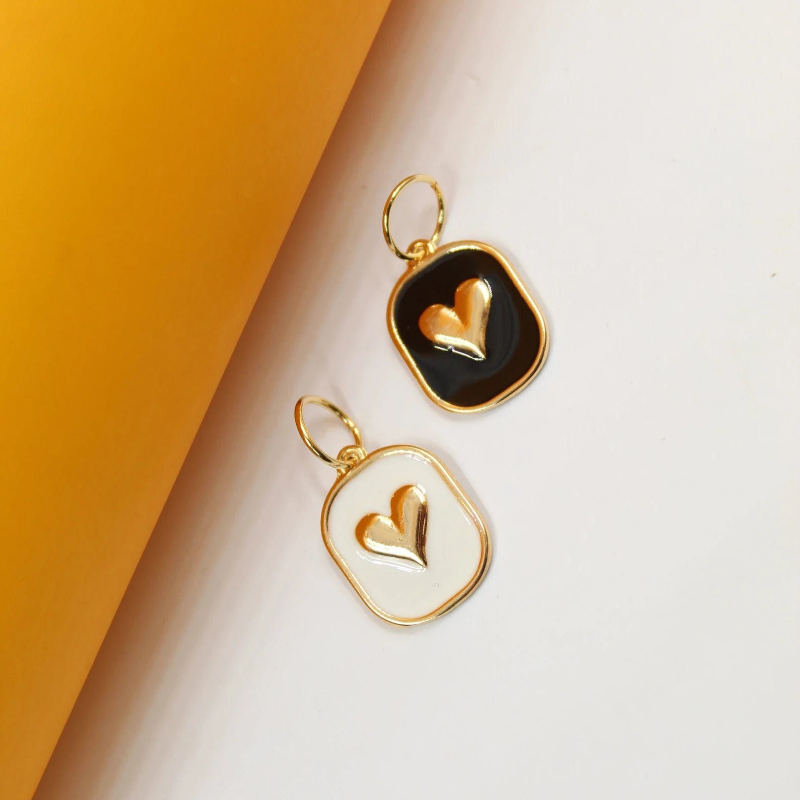 Two gold-trimmed enamel heart charms lie against a white and yellow background, showcasing a black and a white pendant.