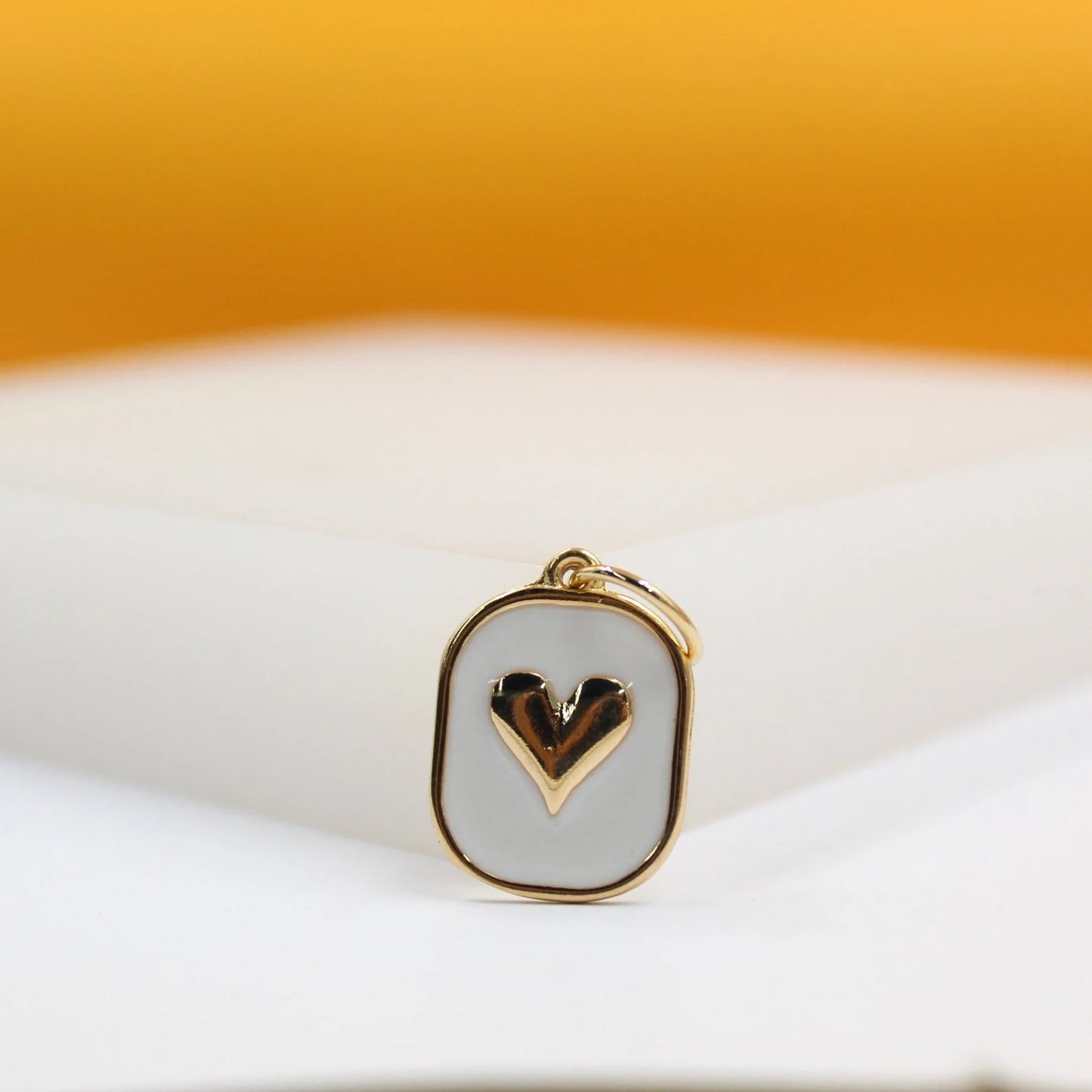 A gold-rimmed rectangular pendant features a white enamel background and a raised gold heart in the center, resting on a white display surface.