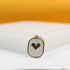 A gold-rimmed rectangular pendant features a white enamel background and a raised gold heart in the center, resting on a white display surface.