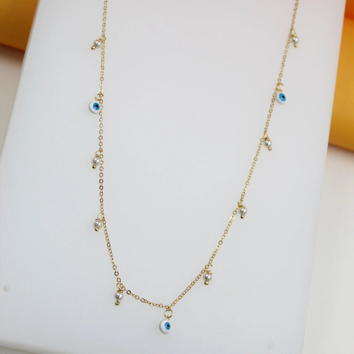 A delicate gold chain necklace drapes against a white surface, featuring dangling evil eye charms and small white pearls.