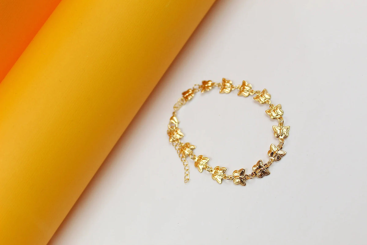 A delicate gold butterfly bracelet rests on a white surface next to a yellow cylinder.