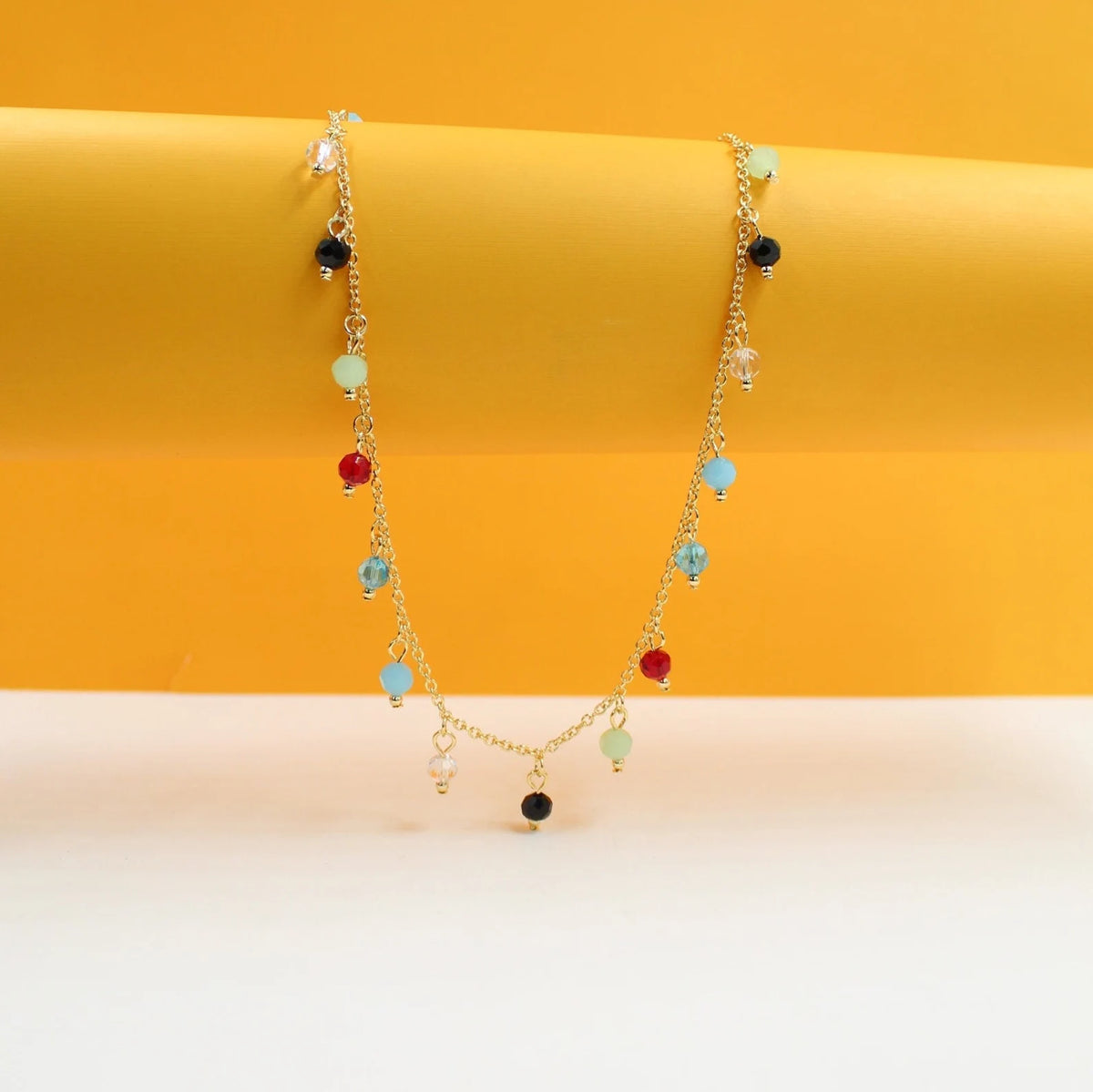 A delicate gold chain necklace drapes against a vibrant yellow and white backdrop, featuring colorful gemstone charms in shades of red, blue, green, and black.