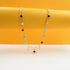 A delicate gold chain necklace drapes against a vibrant yellow and white backdrop, featuring colorful gemstone charms in shades of red, blue, green, and black.