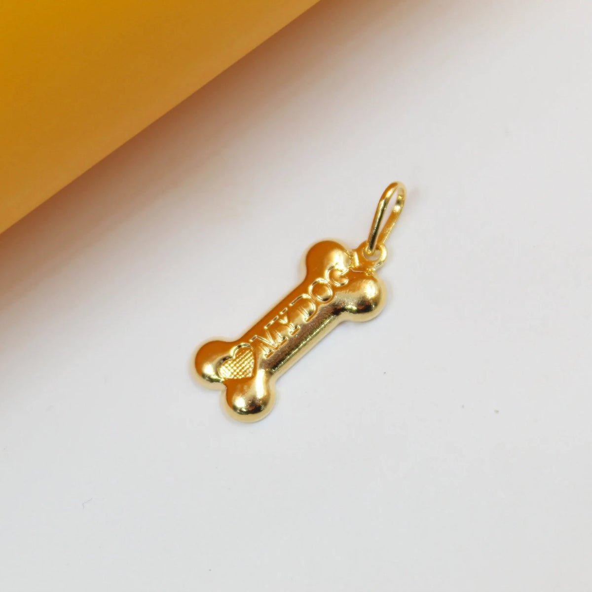 A polished gold bone-shaped pendant rests on a white surface, displaying the engraved phrase "I <3 MY DOG" alongside a small textured heart symbol.
