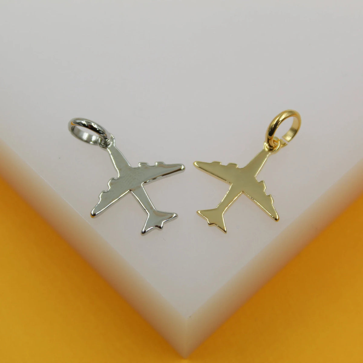 A silver airplane pendant and a gold airplane pendant rest side-by-side on a white display surface against a warm yellow background.
