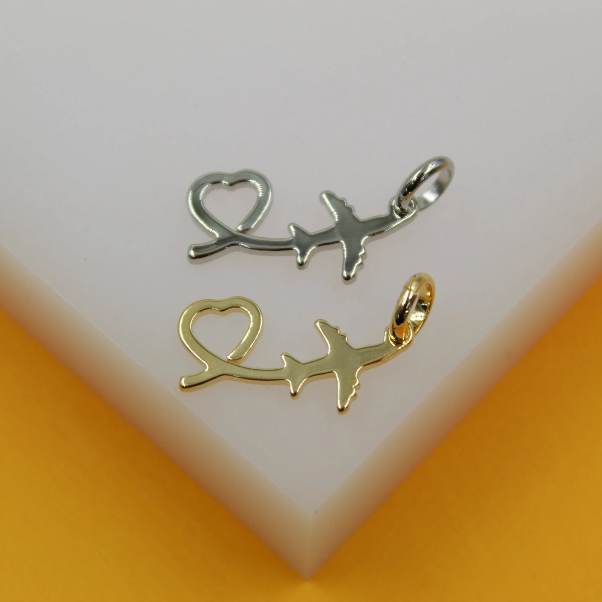 Two airplane and heart travel pendant charms in silver and gold rest on a white display stand against a warm orange background.