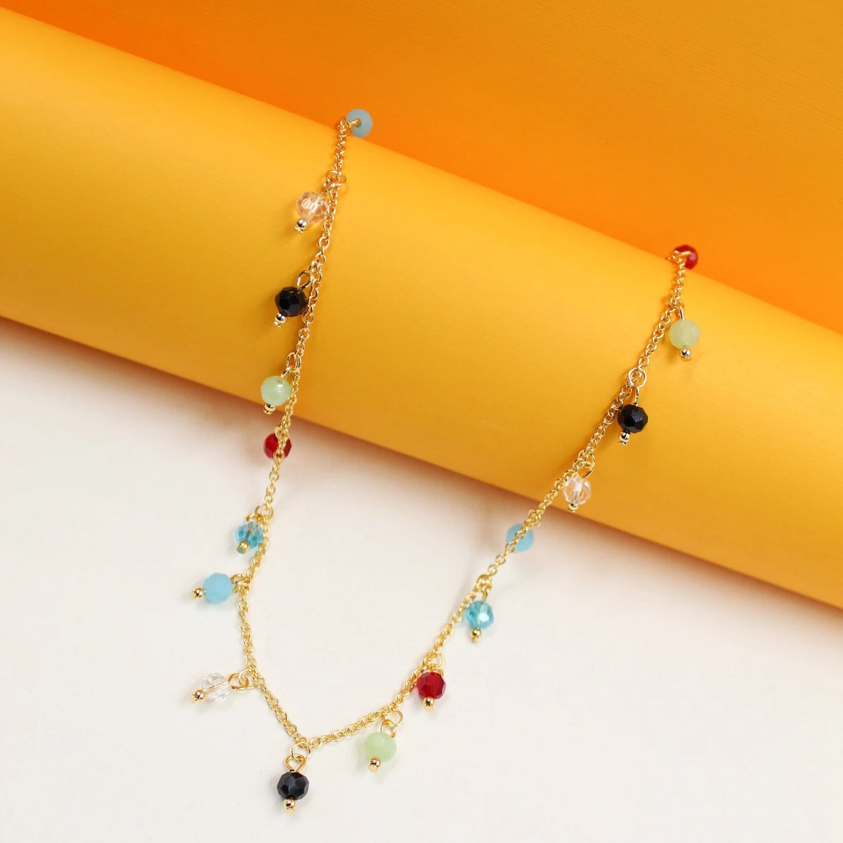 A delicate gold chain necklace with colorful faceted crystal beads drapes across a vibrant yellow and white background.