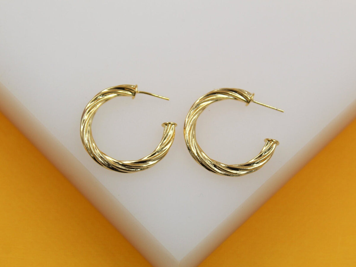 A pair of polished gold twist hoop earrings rests elegantly on a white surface against an orange background.