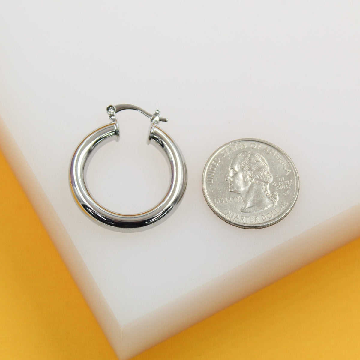 A chunky silver hoop earring rests next to a US quarter coin on a white and yellow display block.