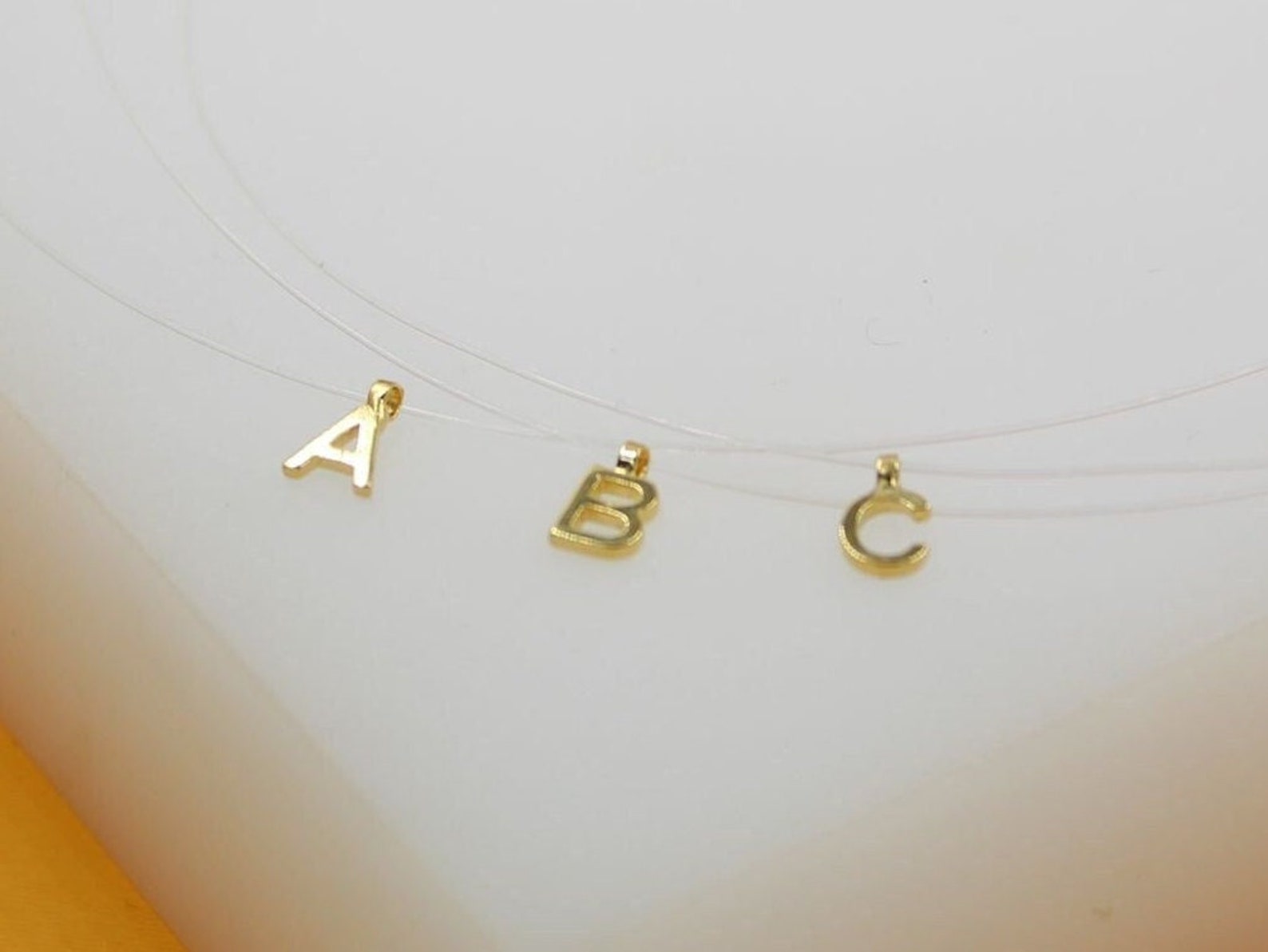 Three delicate transparent necklaces display gold initial letter pendants spelling A, B, and C against a neutral white background.