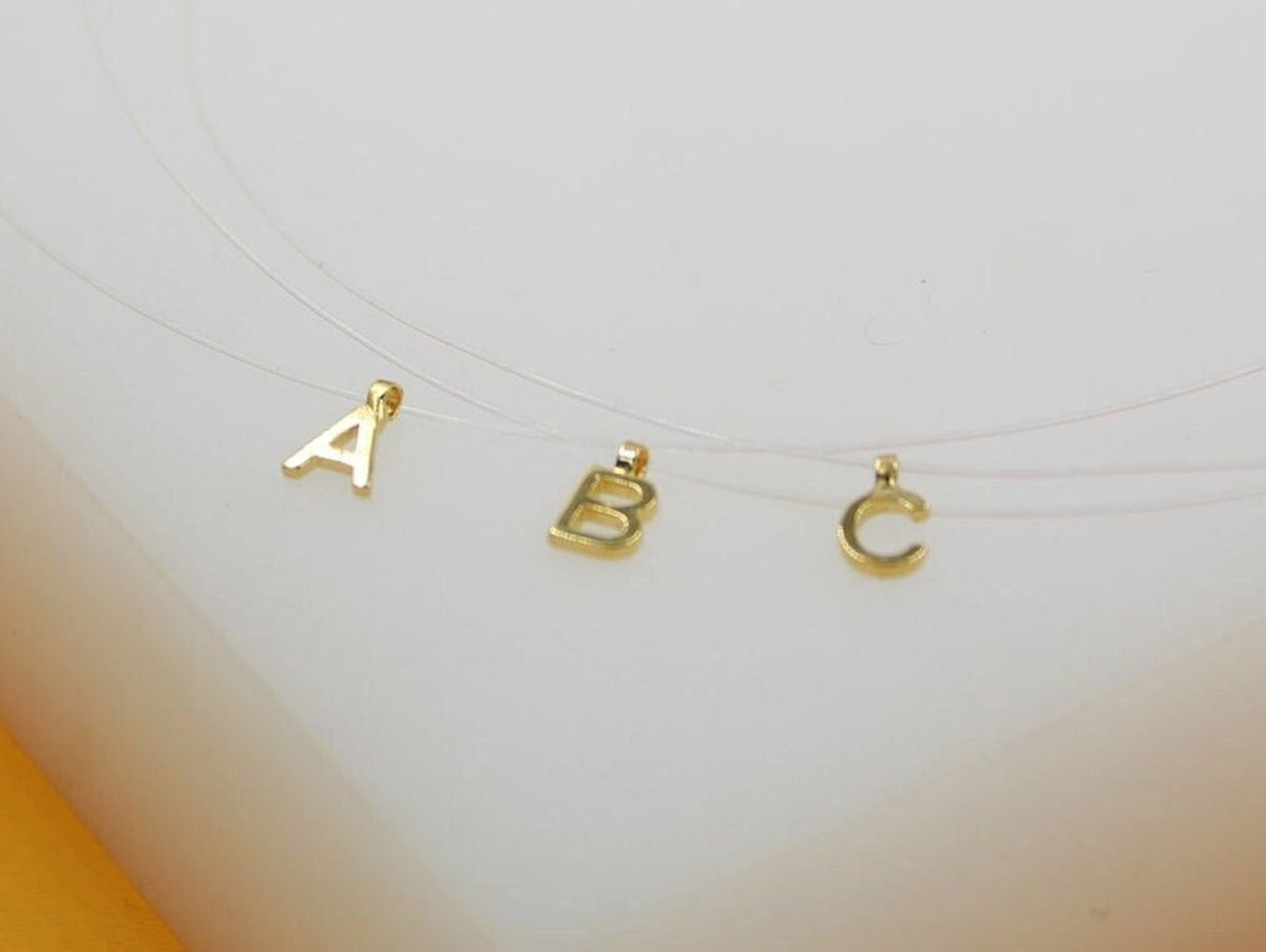 Three delicate gold initial letter charms, A, B, and C, hang on invisible fishing line necklaces against a white background.
