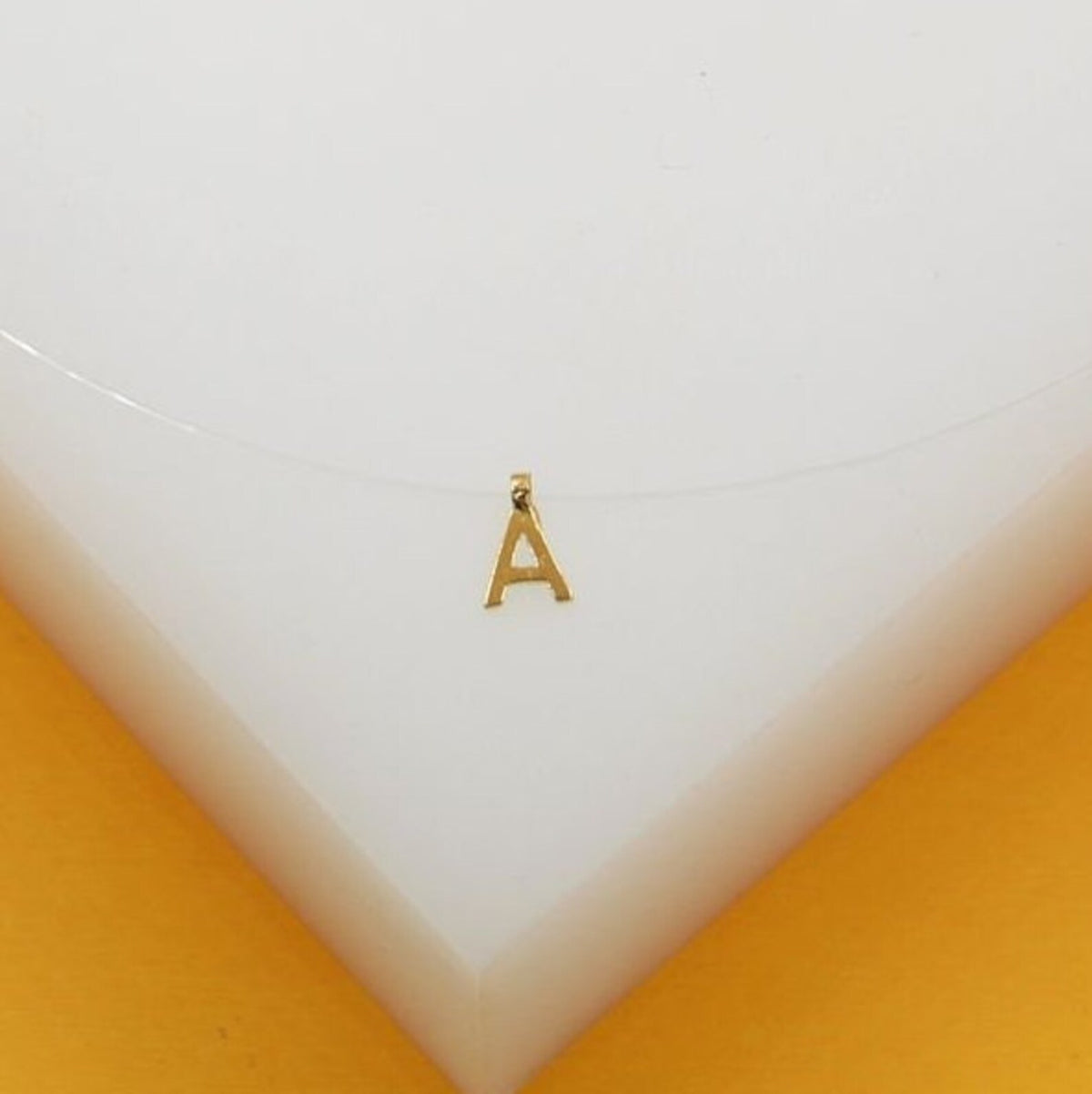 A delicate gold letter A pendant hangs on a transparent fishing line necklace against a white and yellow background.