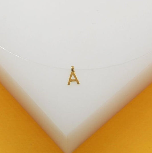 A delicate gold initial A pendant hangs from a thin invisible fishing line necklace resting on a white display stand.