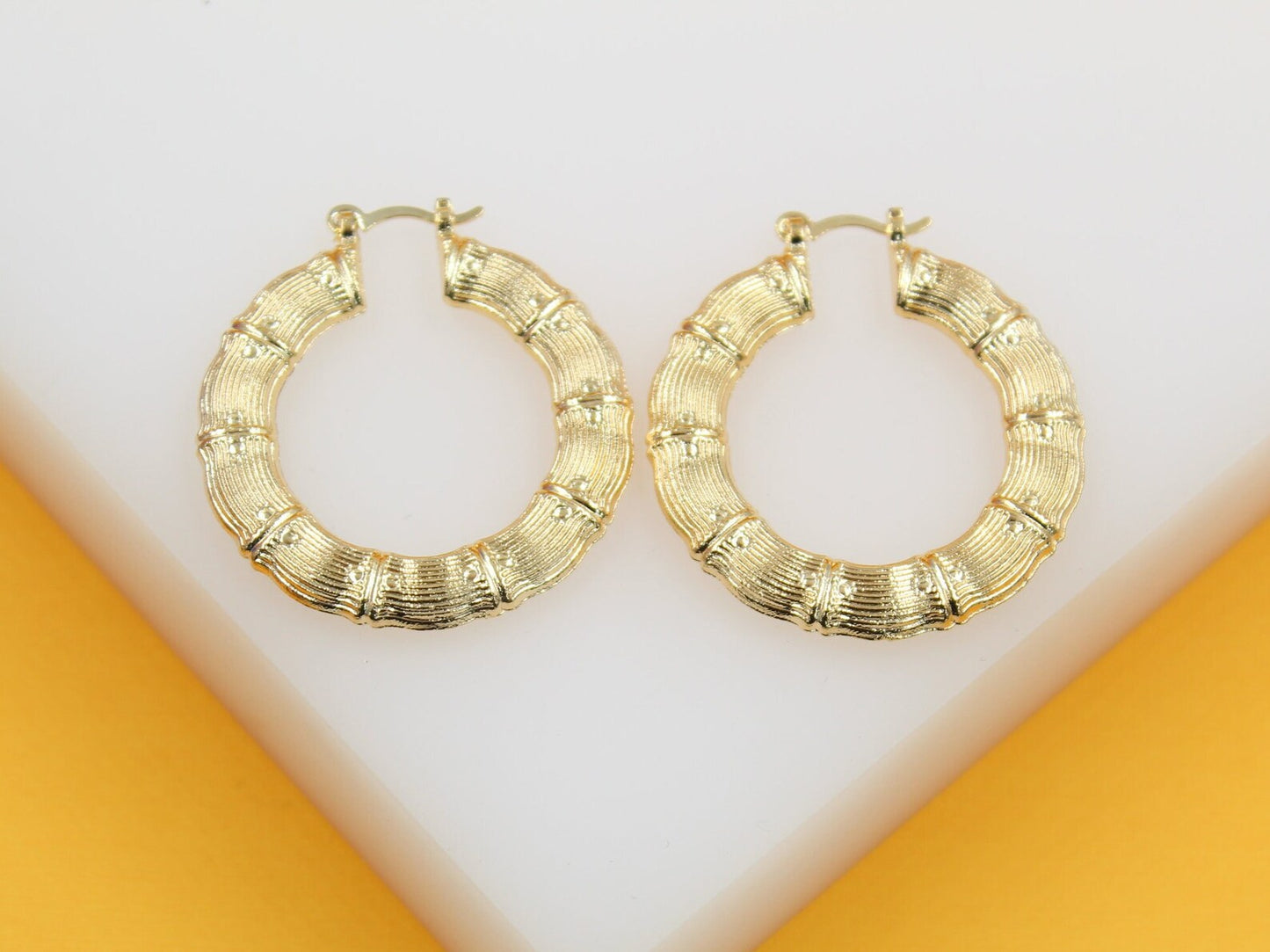 A pair of gold bamboo hoop earrings rests against a white and yellow background.