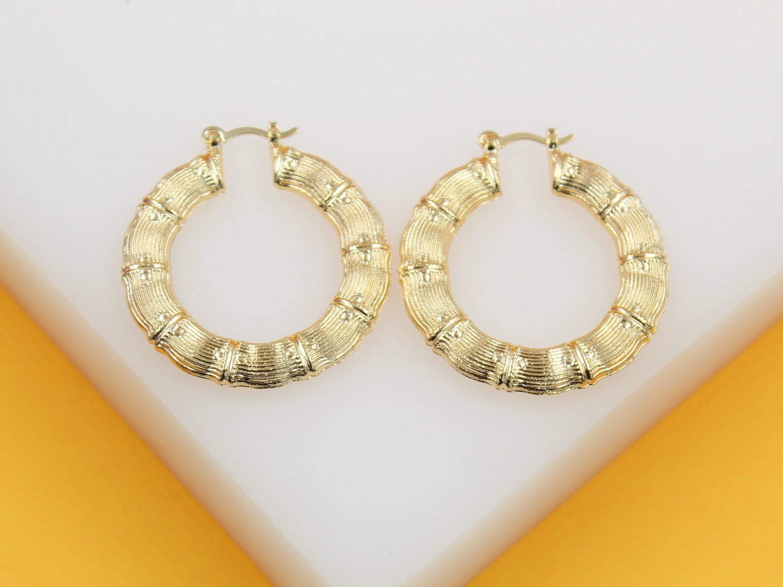 A pair of gold bamboo hoop earrings rests against a white and yellow background.