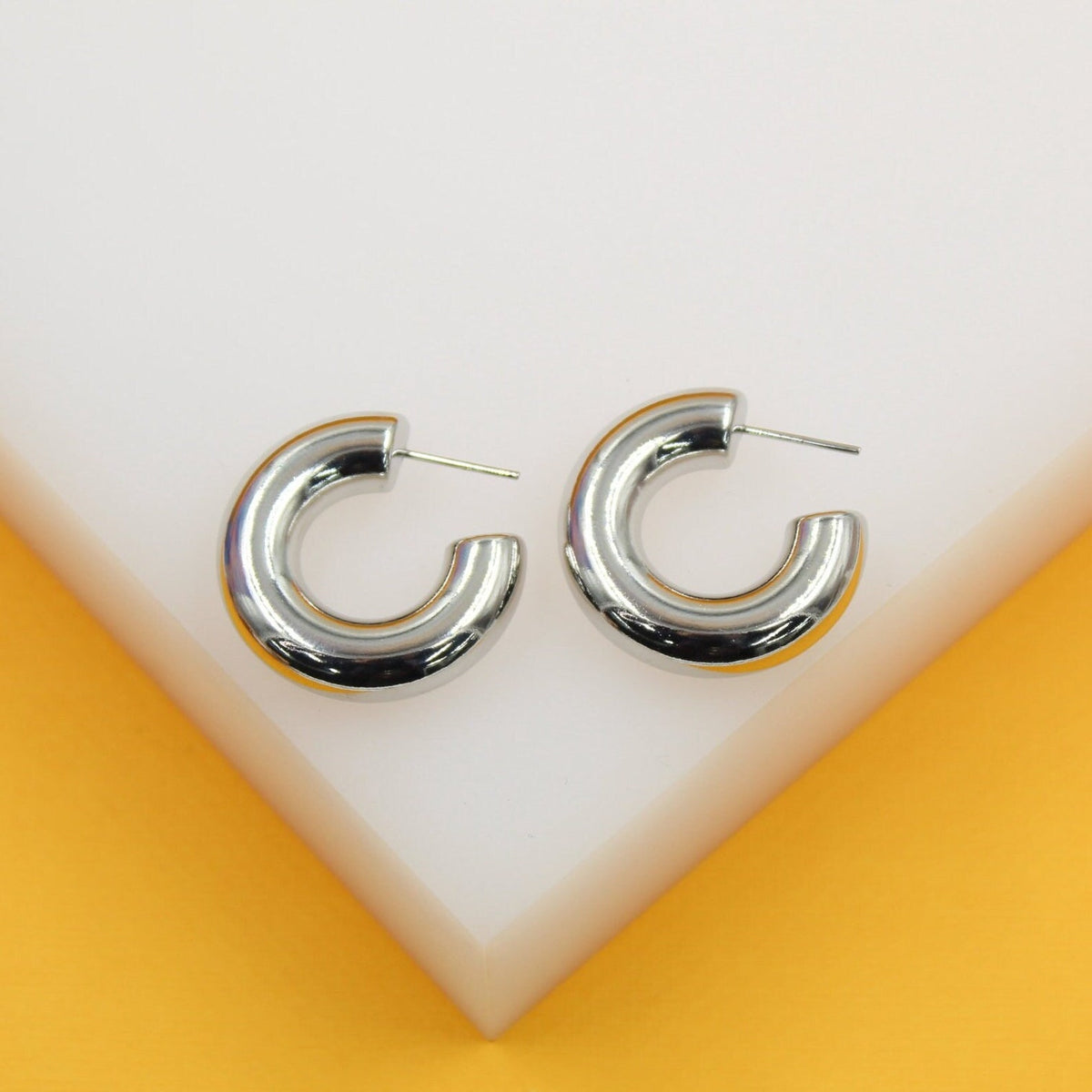 A pair of shiny silver chunky hoop earrings rests on a white display surface against a yellow background.