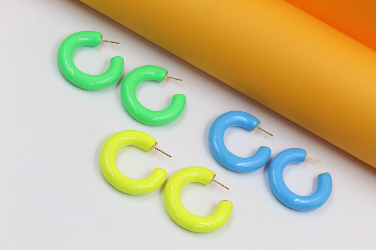 Several colorful, chunky enamel hoop earrings in neon green, yellow, and light blue rest on a white surface next to orange cylinders.