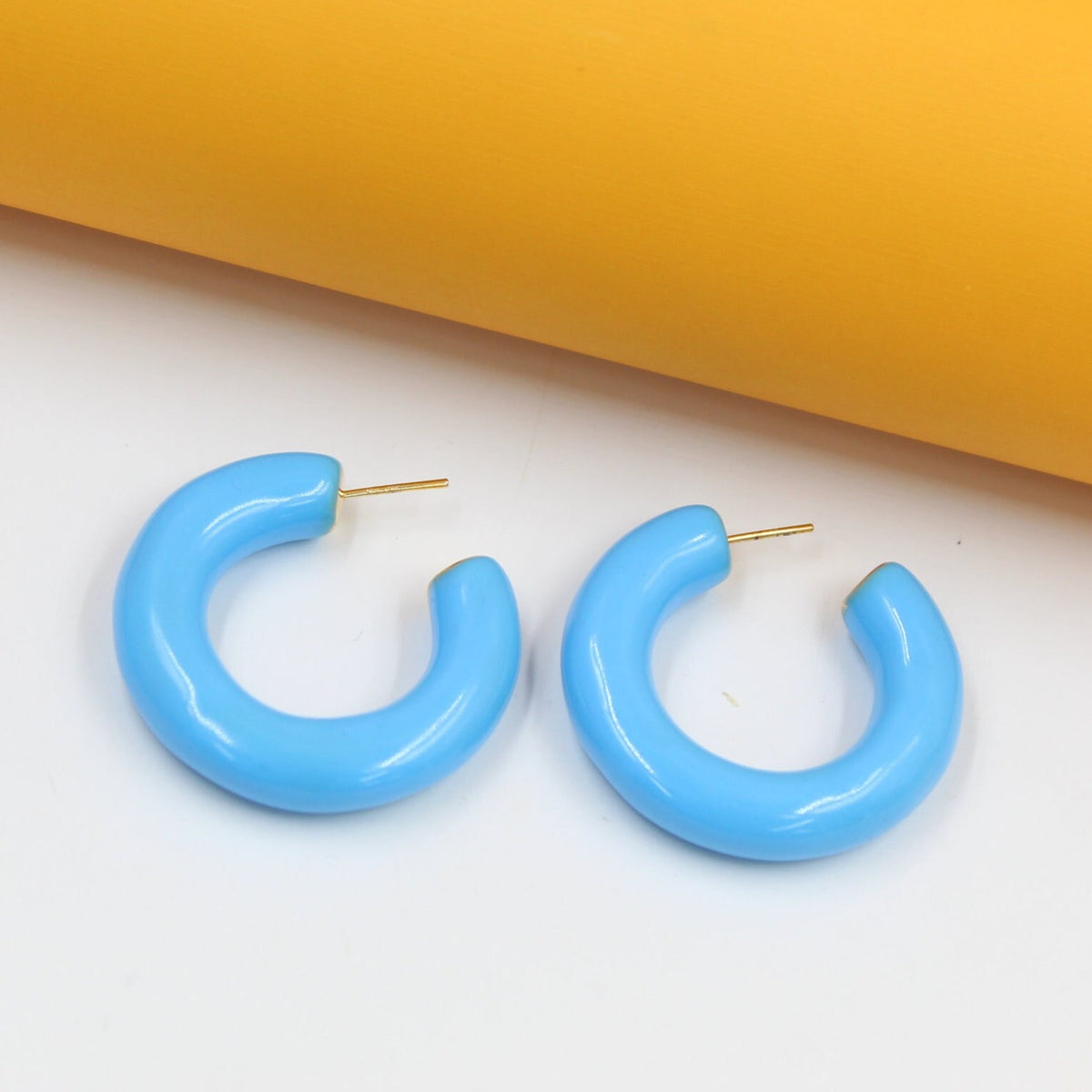 A pair of glossy light blue chunky hoop earrings rests on a white and yellow surface.