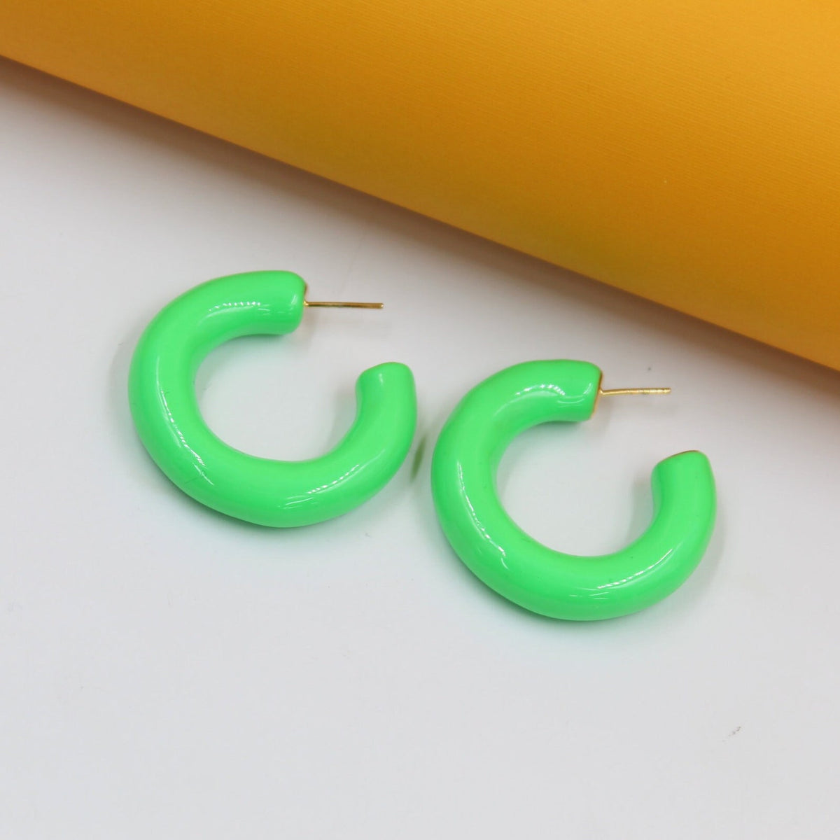 A pair of vibrant neon green enamel hoop earrings rests against a white and yellow background.
