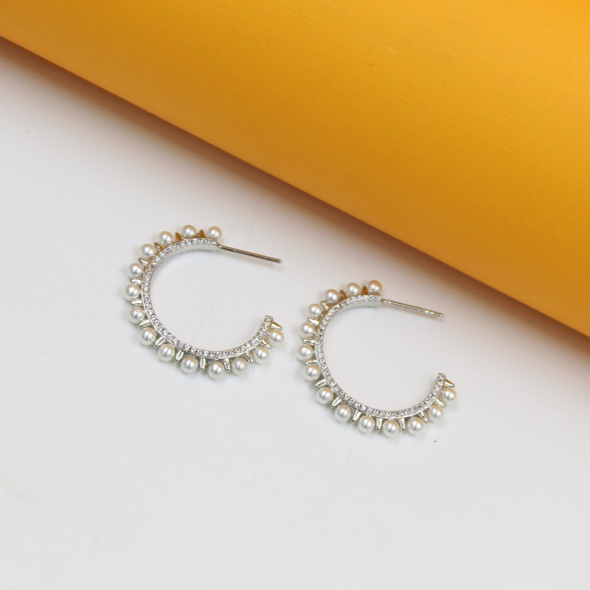 A pair of sterling silver hoop earrings adorned with sparkling cubic zirconia crystals and delicate faux pearls rests against a white and yellow background.