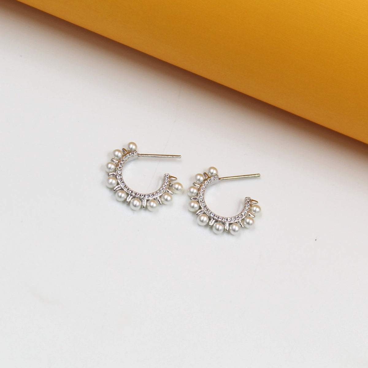 A pair of silver hoop earrings adorned with small pearls and crystals rests on a white surface against an orange backdrop.