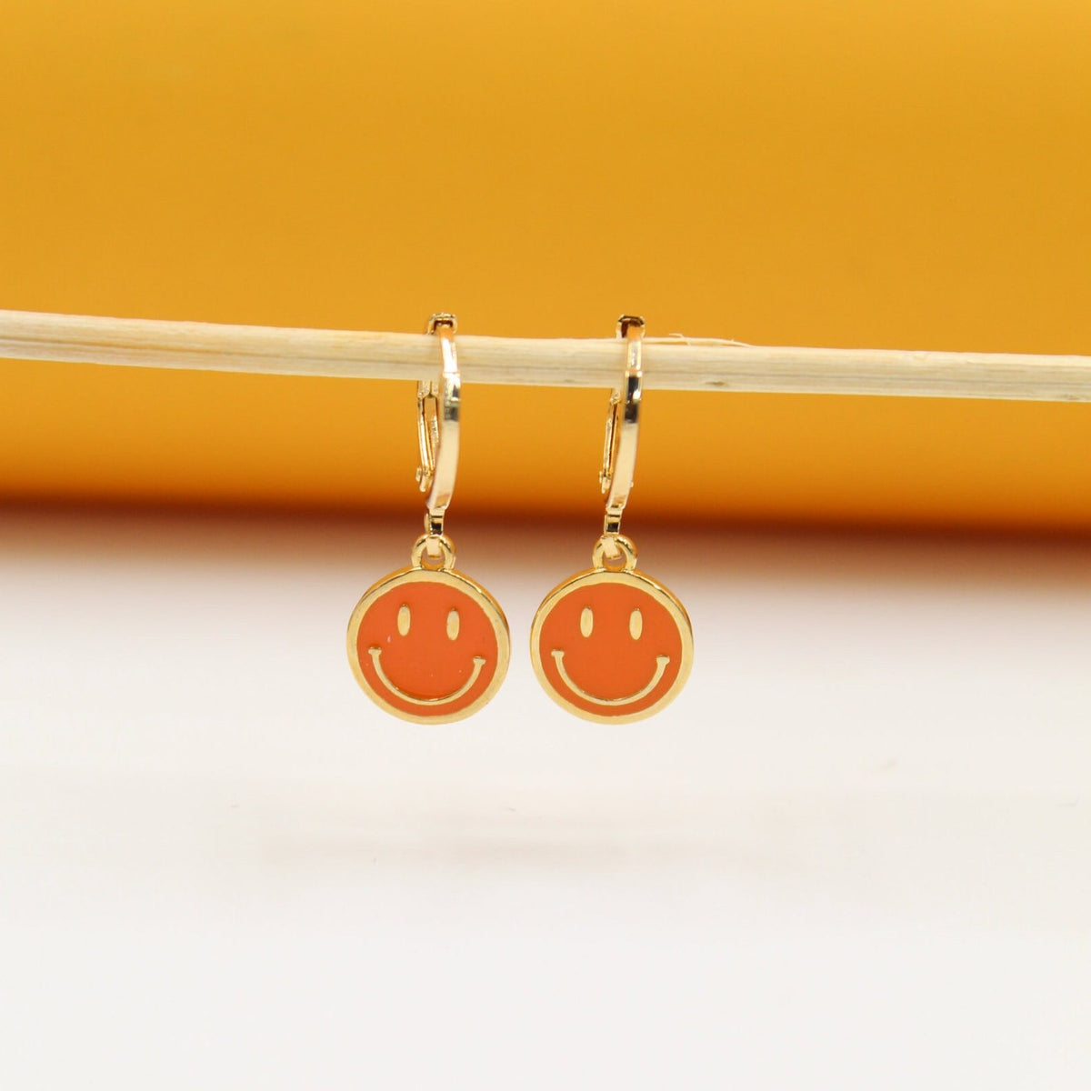 A pair of gold huggie hoop earrings featuring orange enamel smiley face charms hangs from a thin wooden stick against a yellow backdrop.