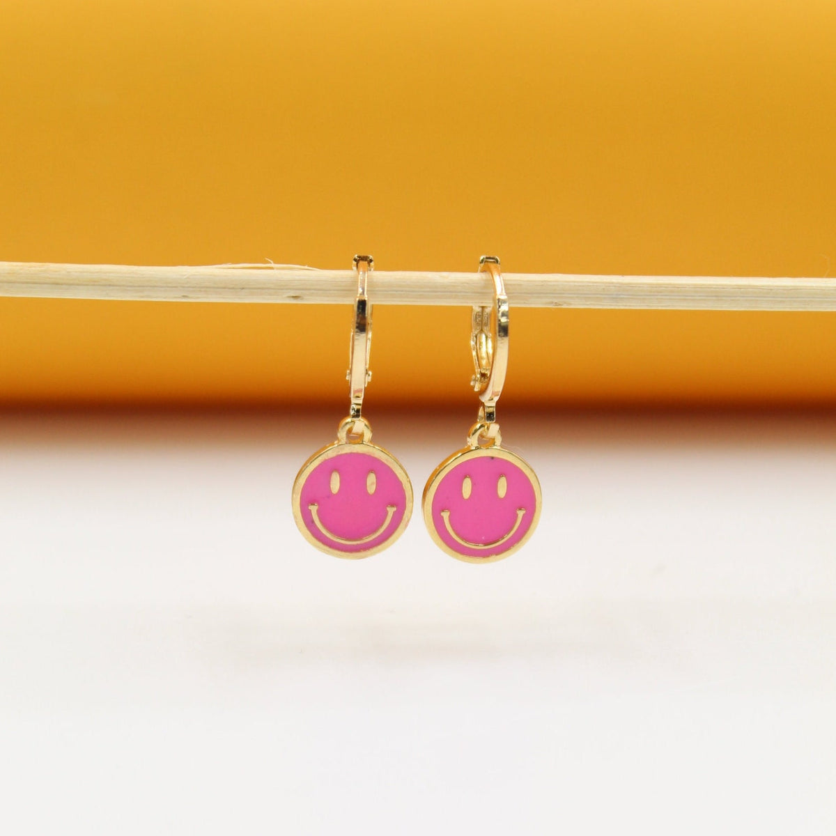 A pair of gold huggie hoop earrings featuring pink enamel smiley face charms hang from a wooden stick against a yellow backdrop.