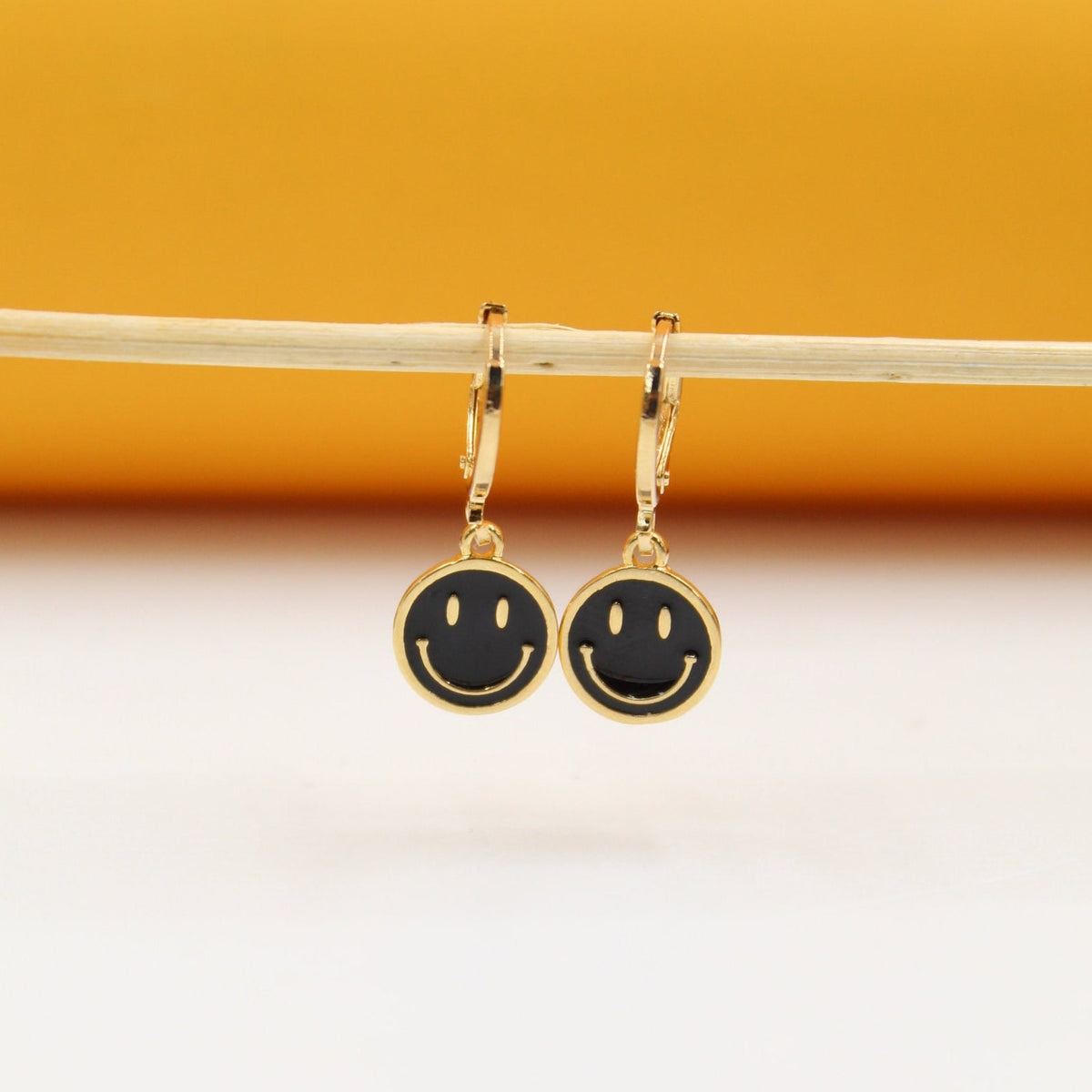 A pair of gold huggie hoop earrings featuring black smiley face charms hangs from a thin wooden stick against an orange and white background.
