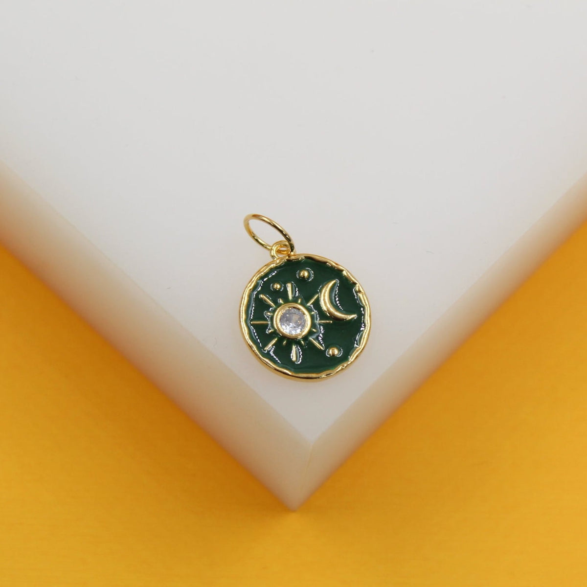 A round gold pendant with green enamel features a sparkling crystal sun and a crescent moon against a white and yellow display background.