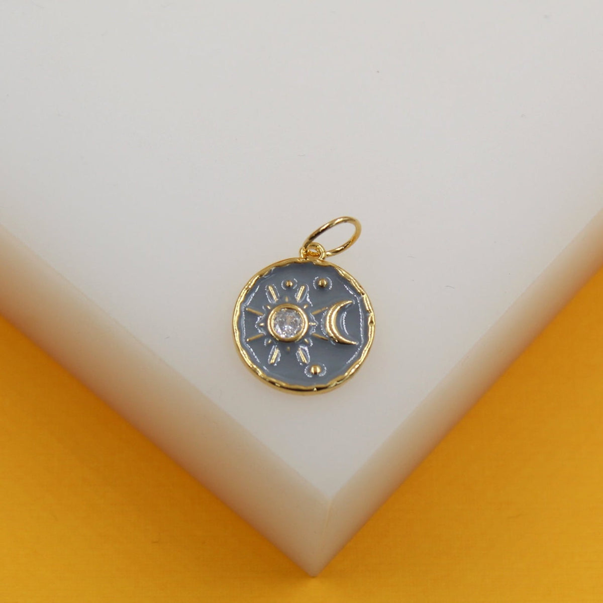 A gold celestial charm featuring a gray enameled background with a rhinestone sun and a crescent moon rests on a white display surface.