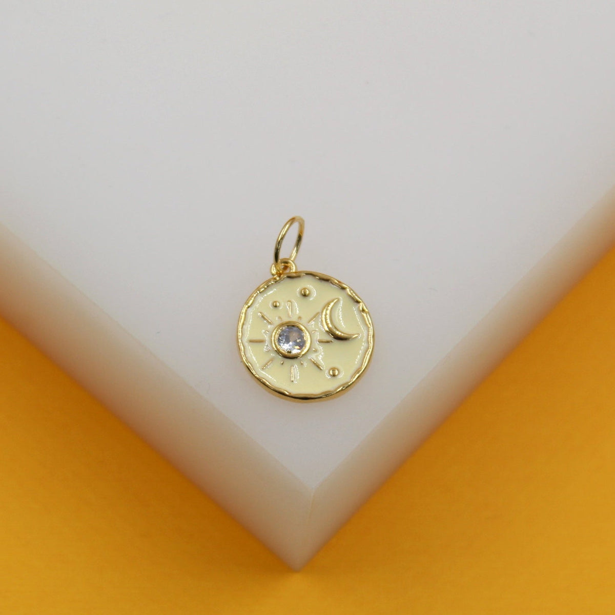 A gold celestial pendant features a sun and moon enamel design with a sparkling center crystal resting on a white display block.
