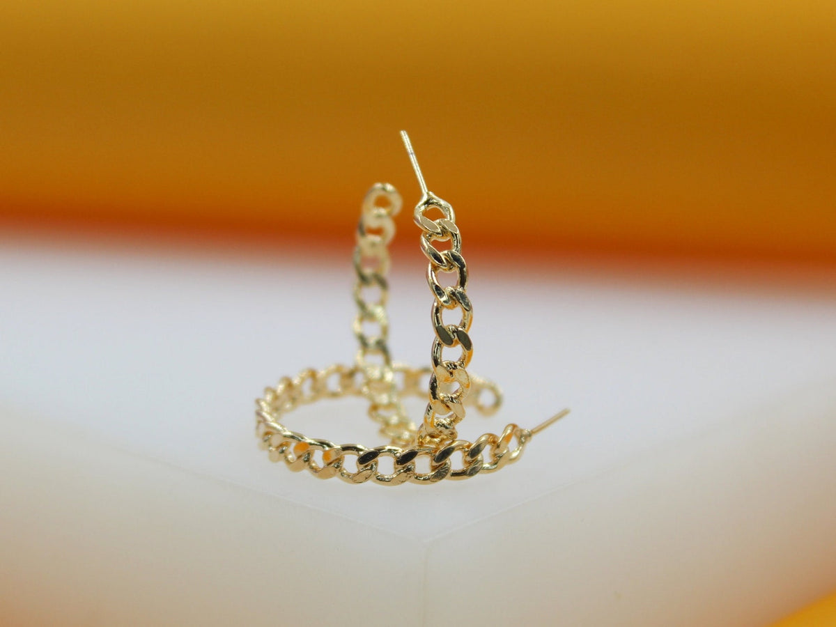 A pair of gold hoop earrings with an intricate chain-link design rests on a white surface against an orange background.