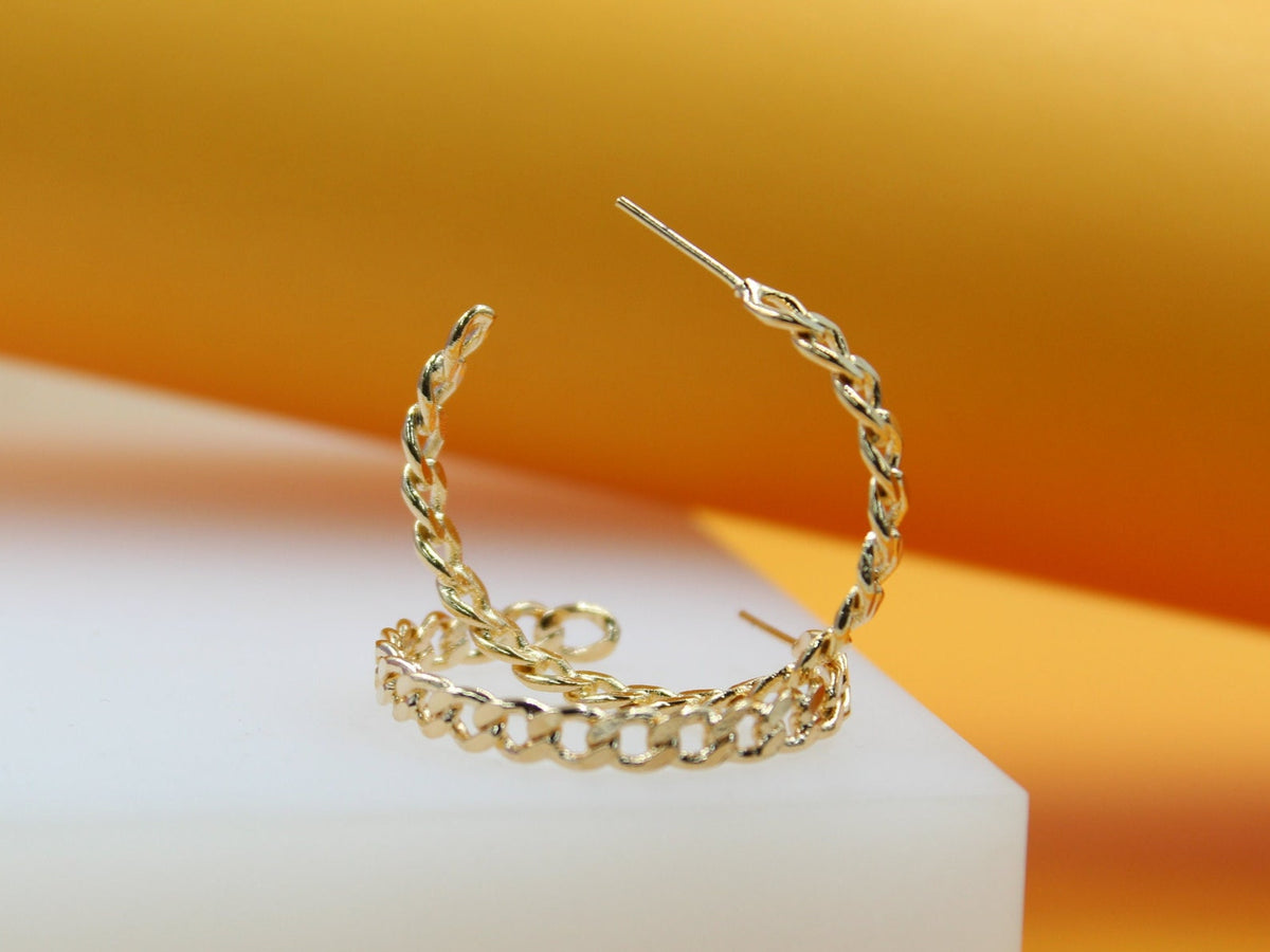 A pair of gold chain-link hoop earrings rests on a white display block against an orange backdrop.