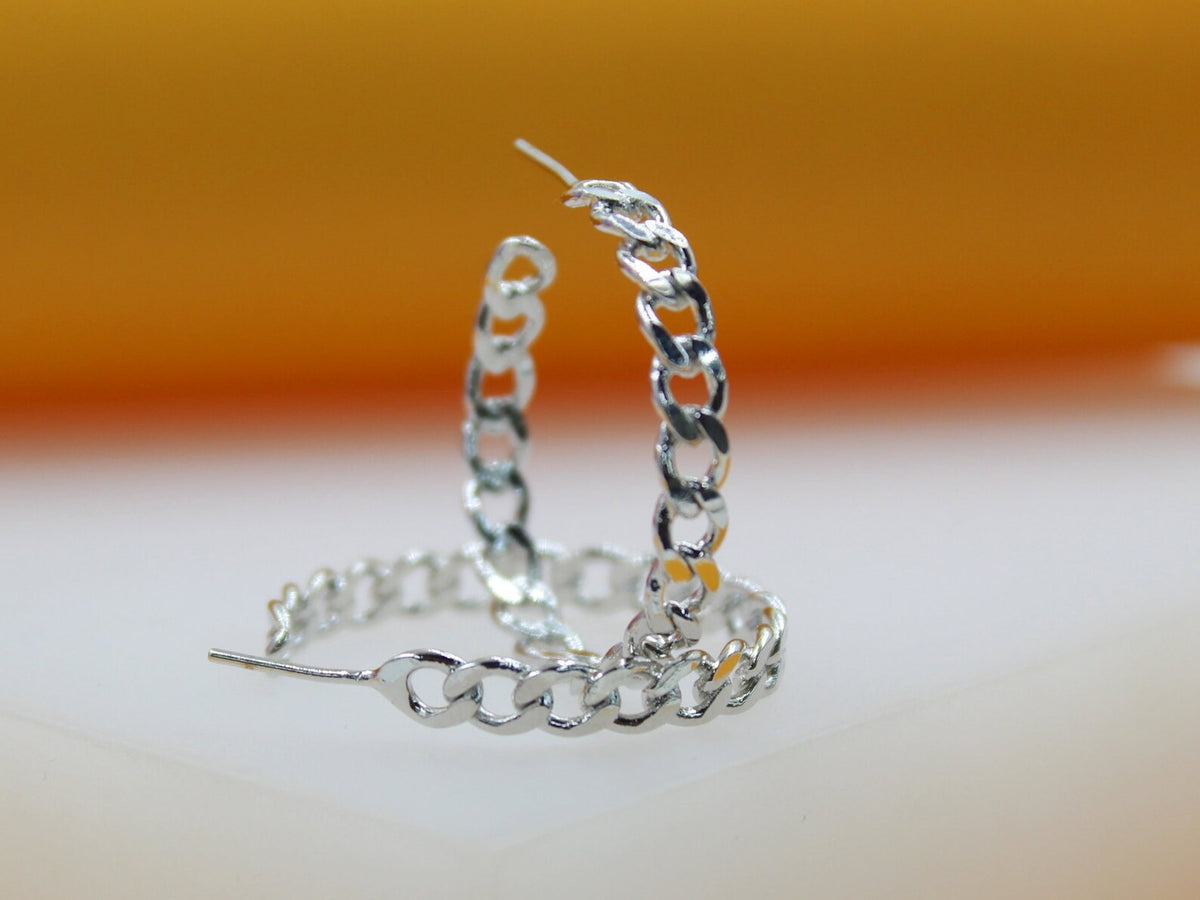 A pair of polished silver cuban link hoop earrings rests on a smooth surface against a warm orange and white backdrop.