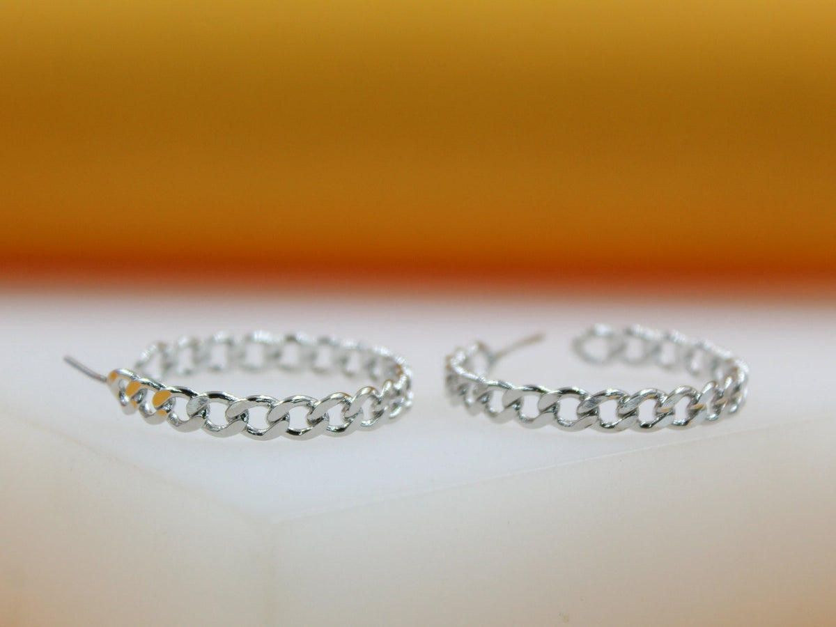 A pair of silver chain-link hoop earrings rests on a smooth surface against a warm orange background.