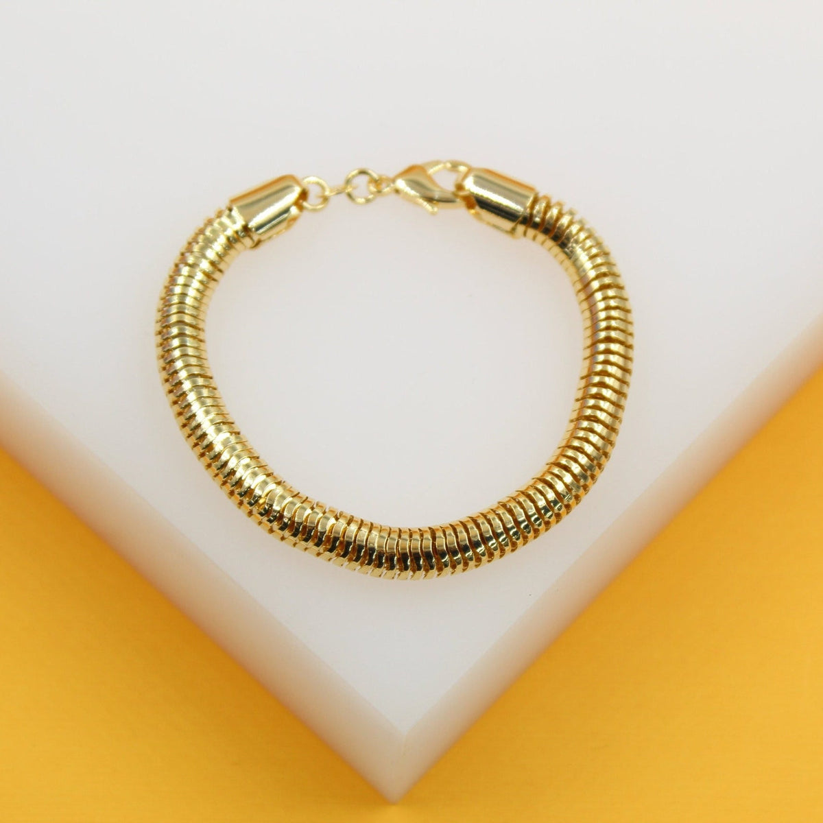 A shiny gold snake chain bracelet rests on a white display surface against a vibrant yellow background.