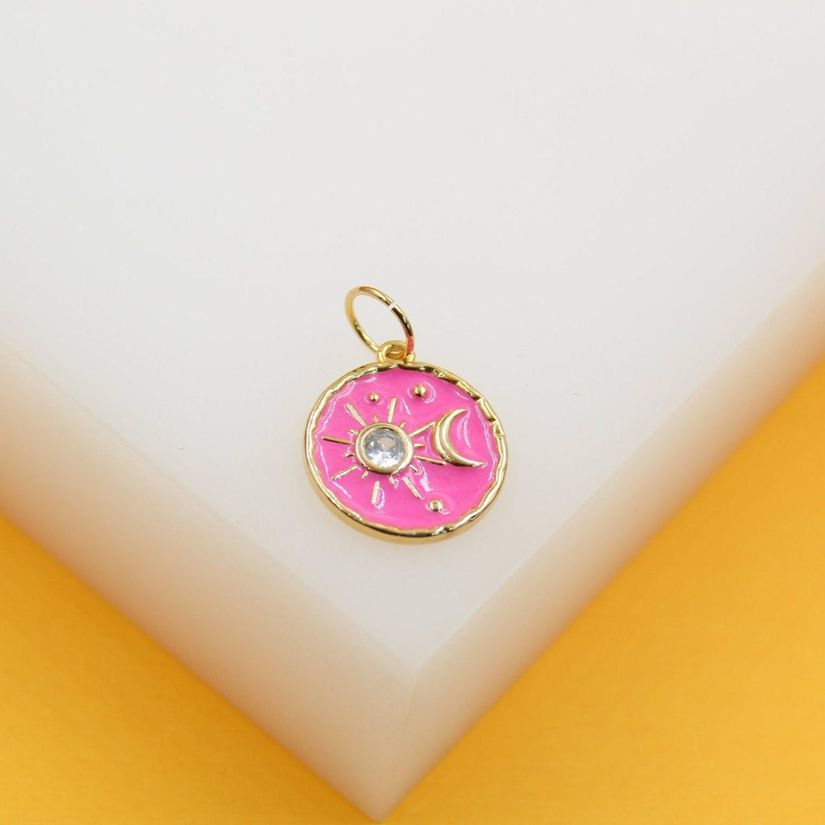 A round gold pendant featuring a vibrant pink enamel background with a central sparkly rhinestone sun and crescent moon rests on a white display block.