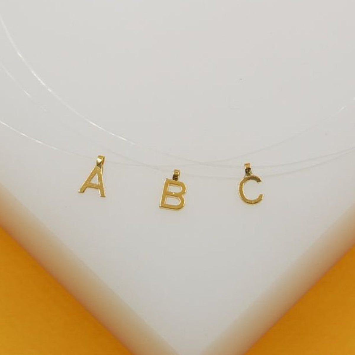 Three gold initial pendants shaped like letters A, B, and C hang delicately from a clear fishing line necklace.