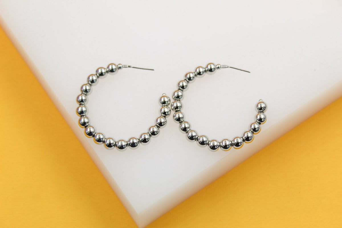 Two silver beaded hoop earrings rest on a white display surface against a vibrant yellow background.